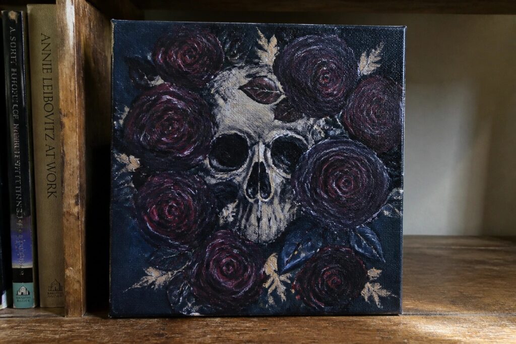 20 Moody Goth Canvas Painting Ideas Built Around Dark Florals Skull with dark roses on black canvas painting atop wooden table with jar nearby