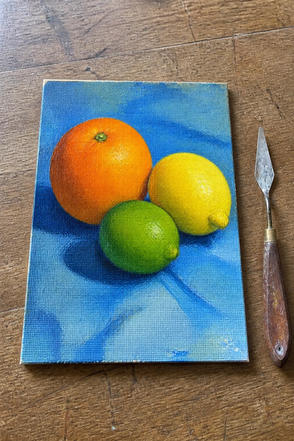 A small oil painting of an orange, a lemon, and a lime on a blue surface with a blue background, placed on a wooden table next to a palette knife.