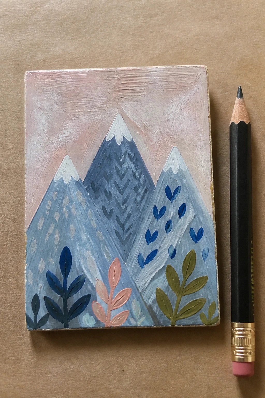 Small blue mountain painting with foliage beside black pencil on brown surface.