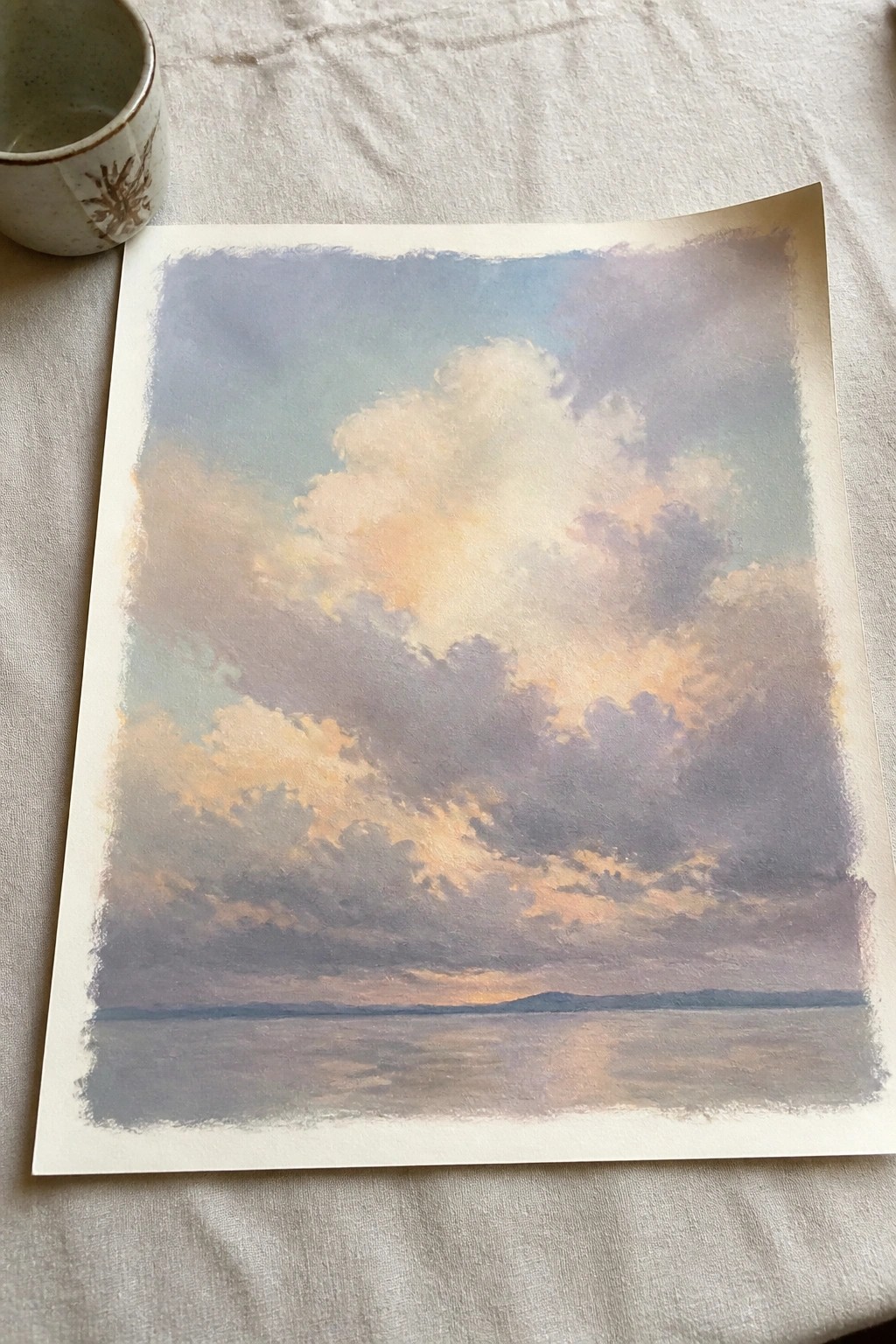 Watercolor painting of colorful clouds over water with ceramic cup on white cloth