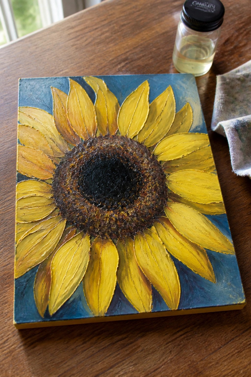 Textured sunflower painting with yellow petals on blue beside glass jar