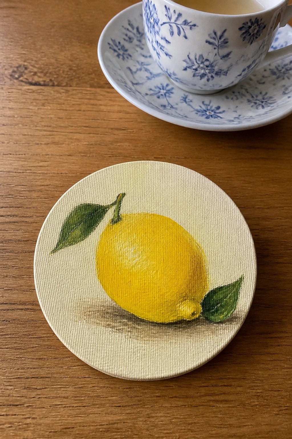 Yellow lemon painted on round beige coaster beside blue floral teacup on wood table