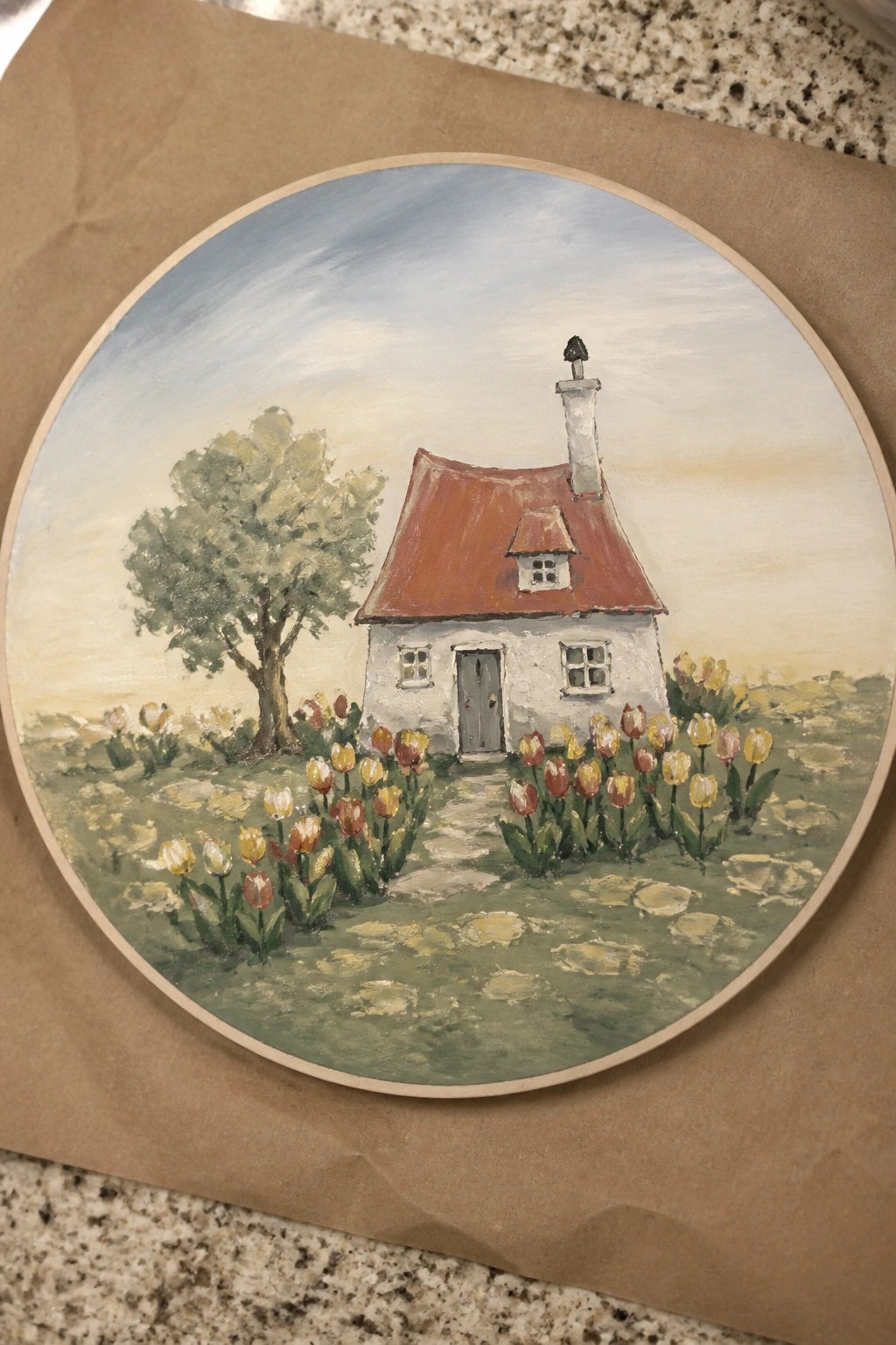 Oval painting of white cottage with red roof amid yellow and red tulips in meadow