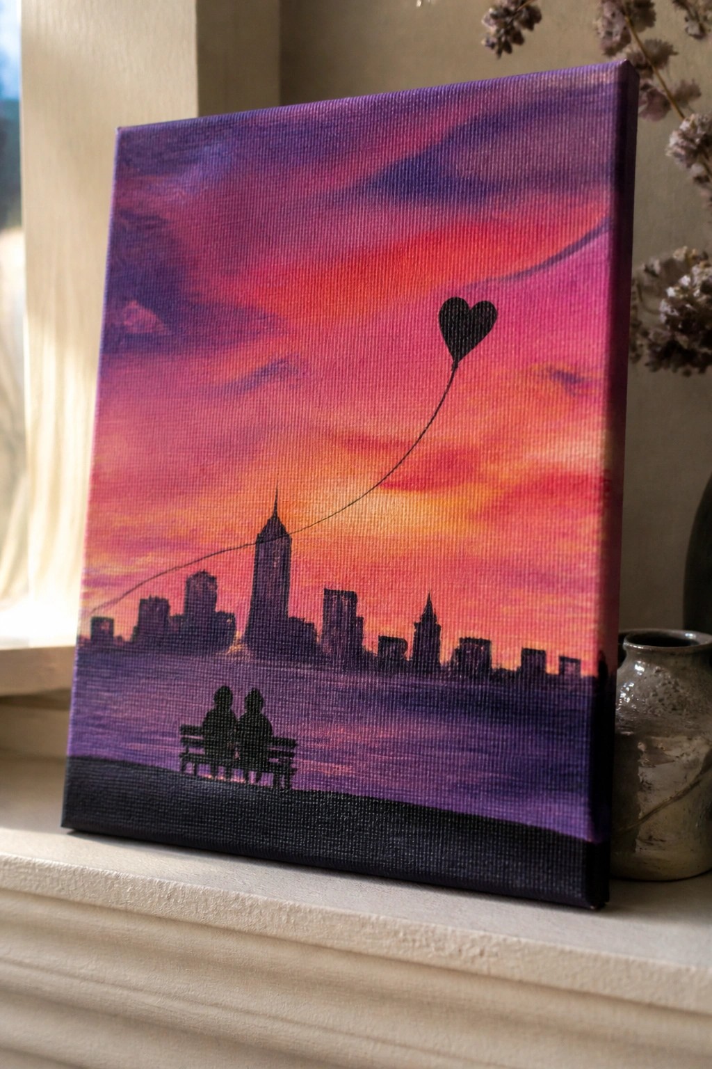 A canvas painting of a sunset skyline with purple and orange clouds, a silhouetted city, two figures on a bench, and a heart balloon on a string.
