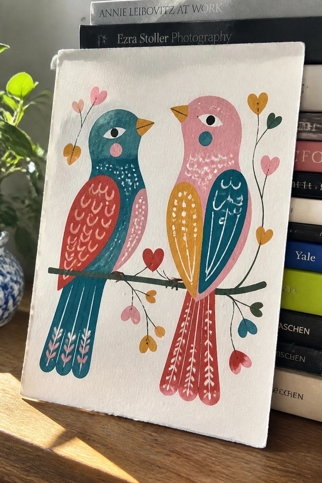 A painting of two colorful stylized birds facing each other on a thin branch with small hearts scattered around them.