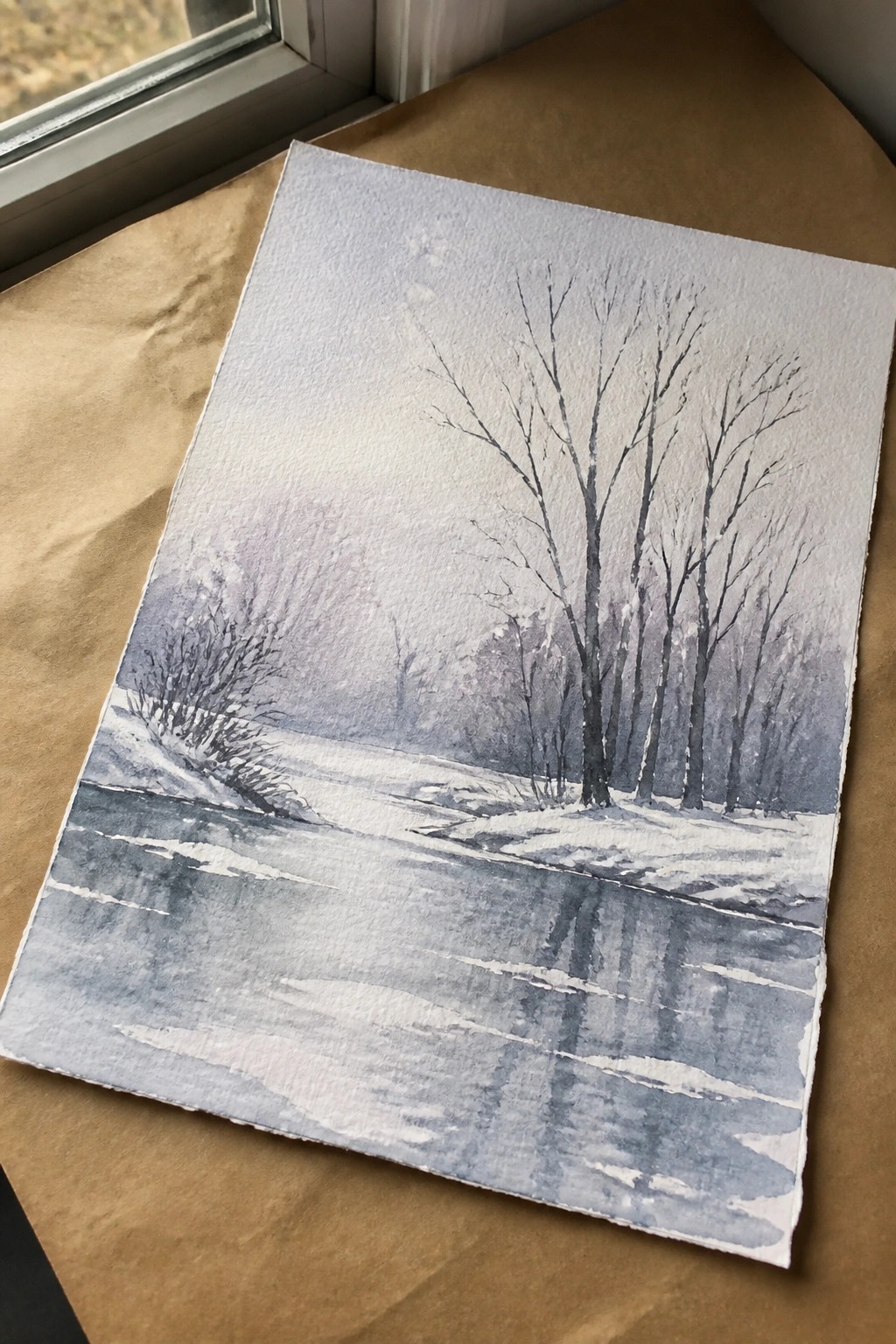 Watercolor painting of snowy winter landscape with bare trees on brown paper by window