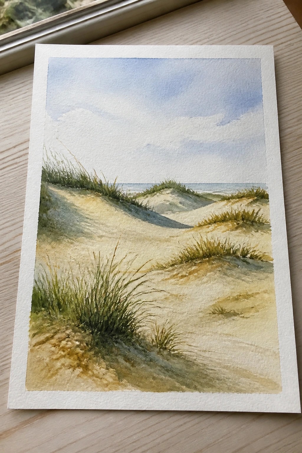 Watercolor landscape of grassy sand dunes leading to ocean under cloudy blue sky