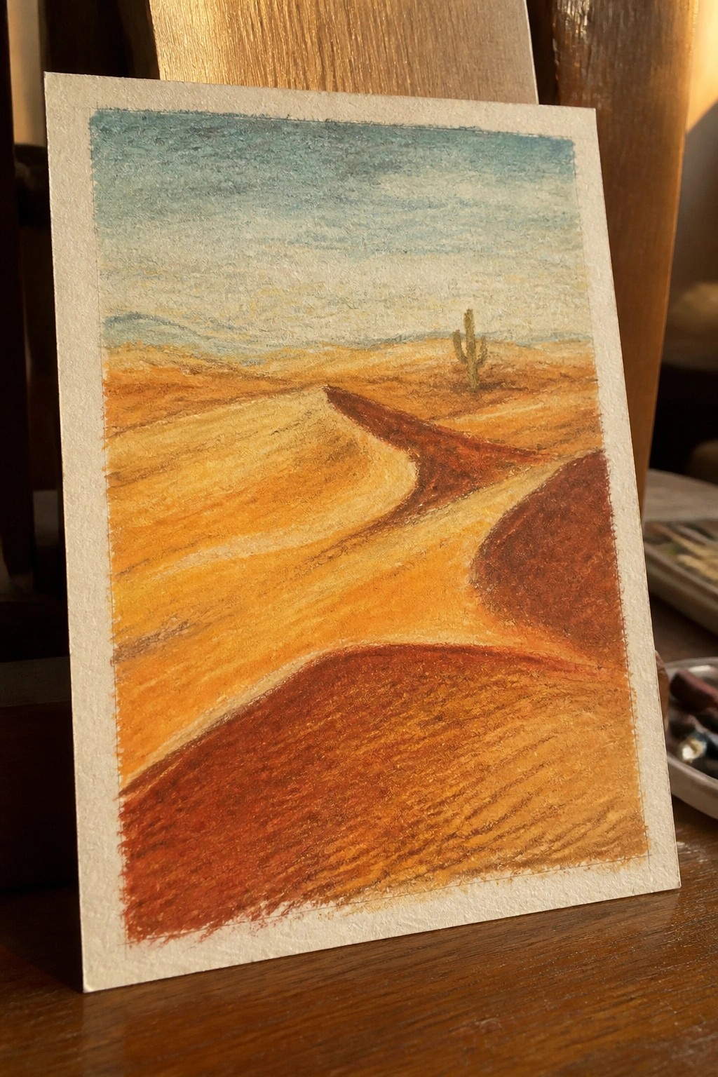 Pastel drawing of orange desert dunes with a lone cactus under blue sky