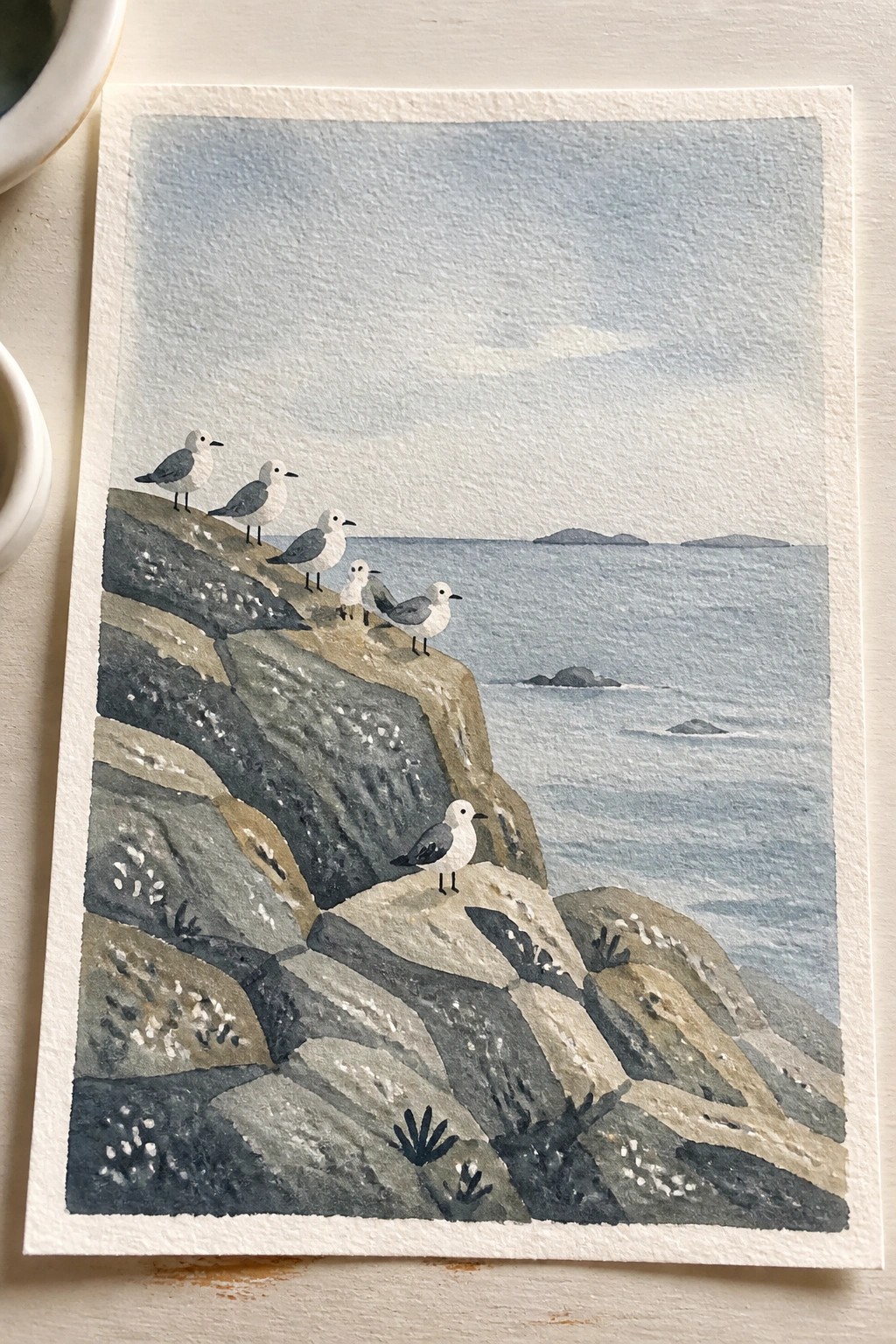 Watercolor seascape with several seagulls perched on layered gray cliffs above blue ocean.