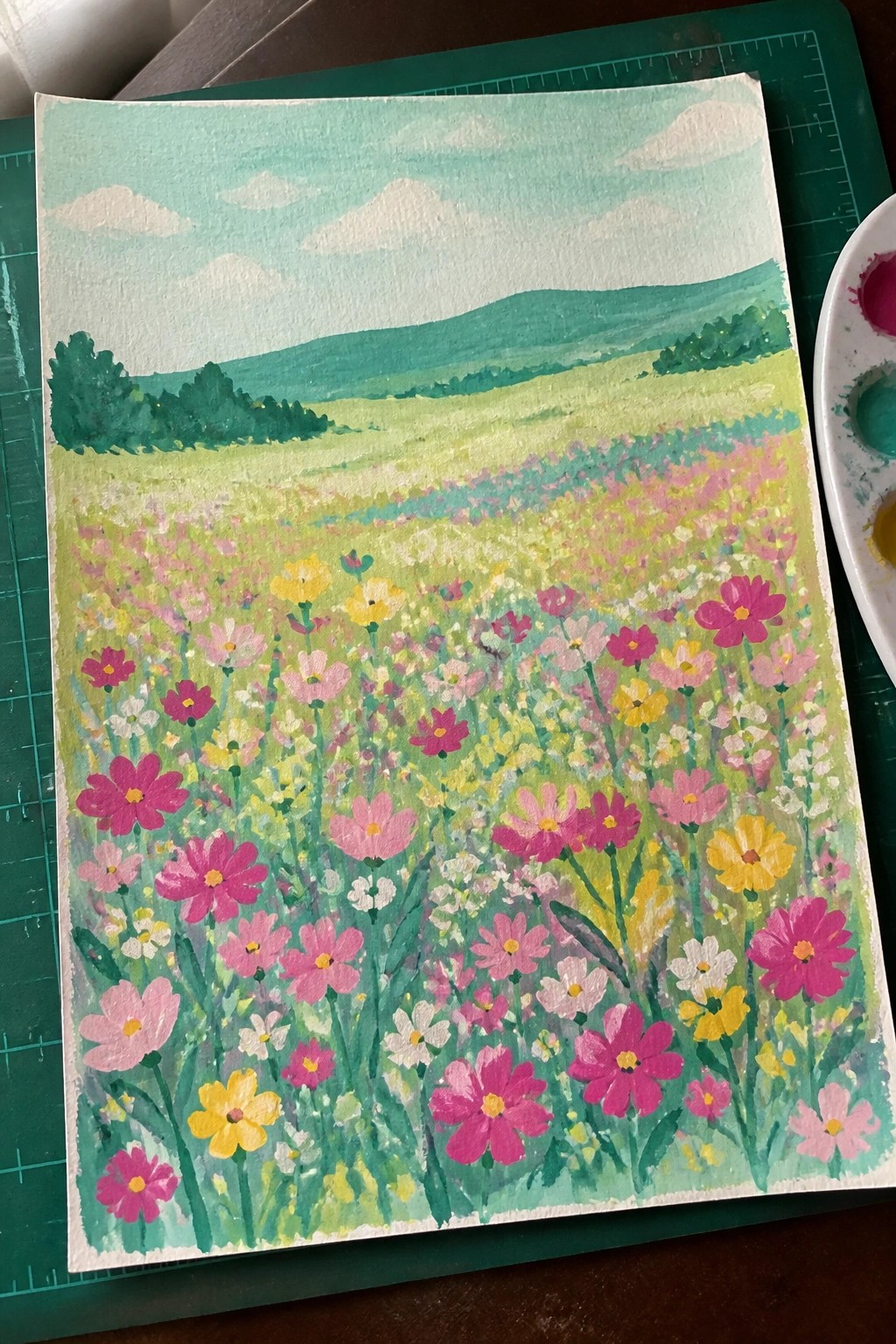 Colorful watercolor painting of a pink and yellow wildflower meadow with green hills and blue sky.