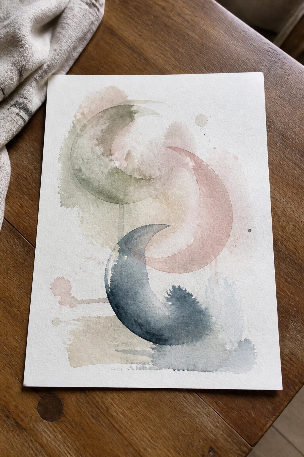 Watercolor painting of overlapping crescent moons in soft green, pink, and blue on paper