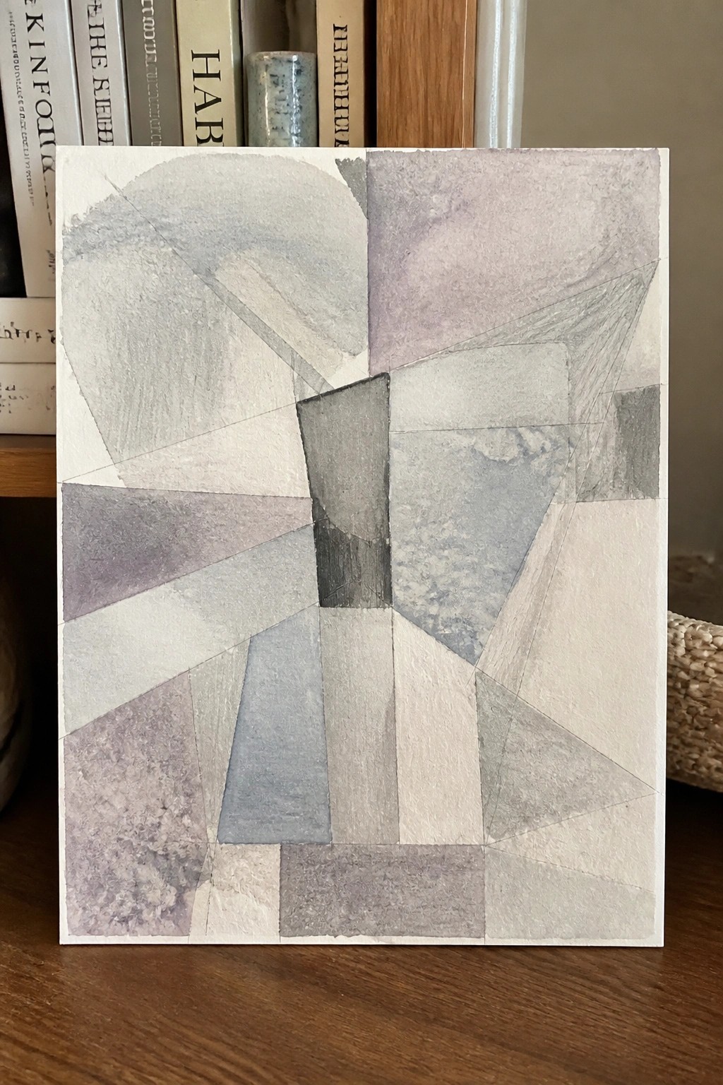 Abstract geometric watercolor painting with muted gray, purple, and beige shapes on wooden surface