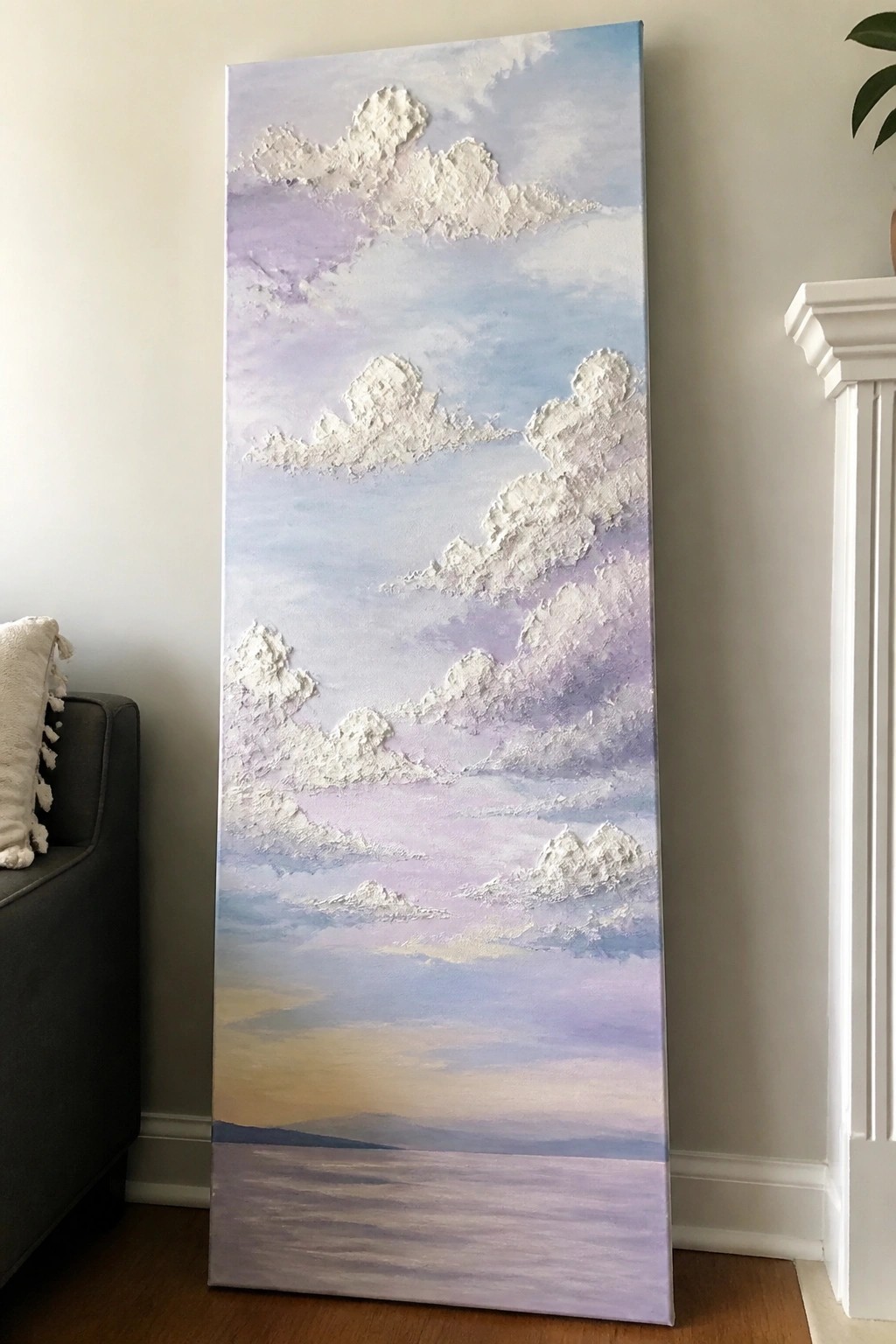 A tall vertical canvas painting of a sky with raised white textured clouds against pastel blue and purple tones, fading to a horizon with water below.