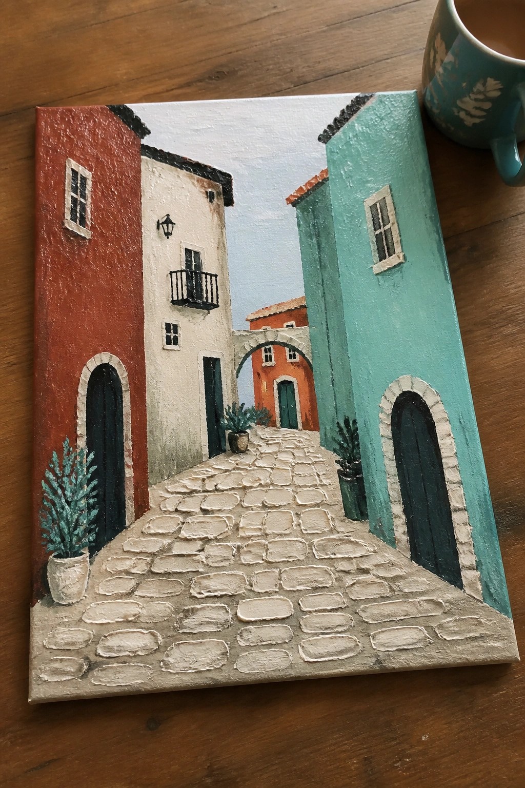 Vibrant painting of cobblestone alley with red, white, and turquoise buildings beside teal mug.