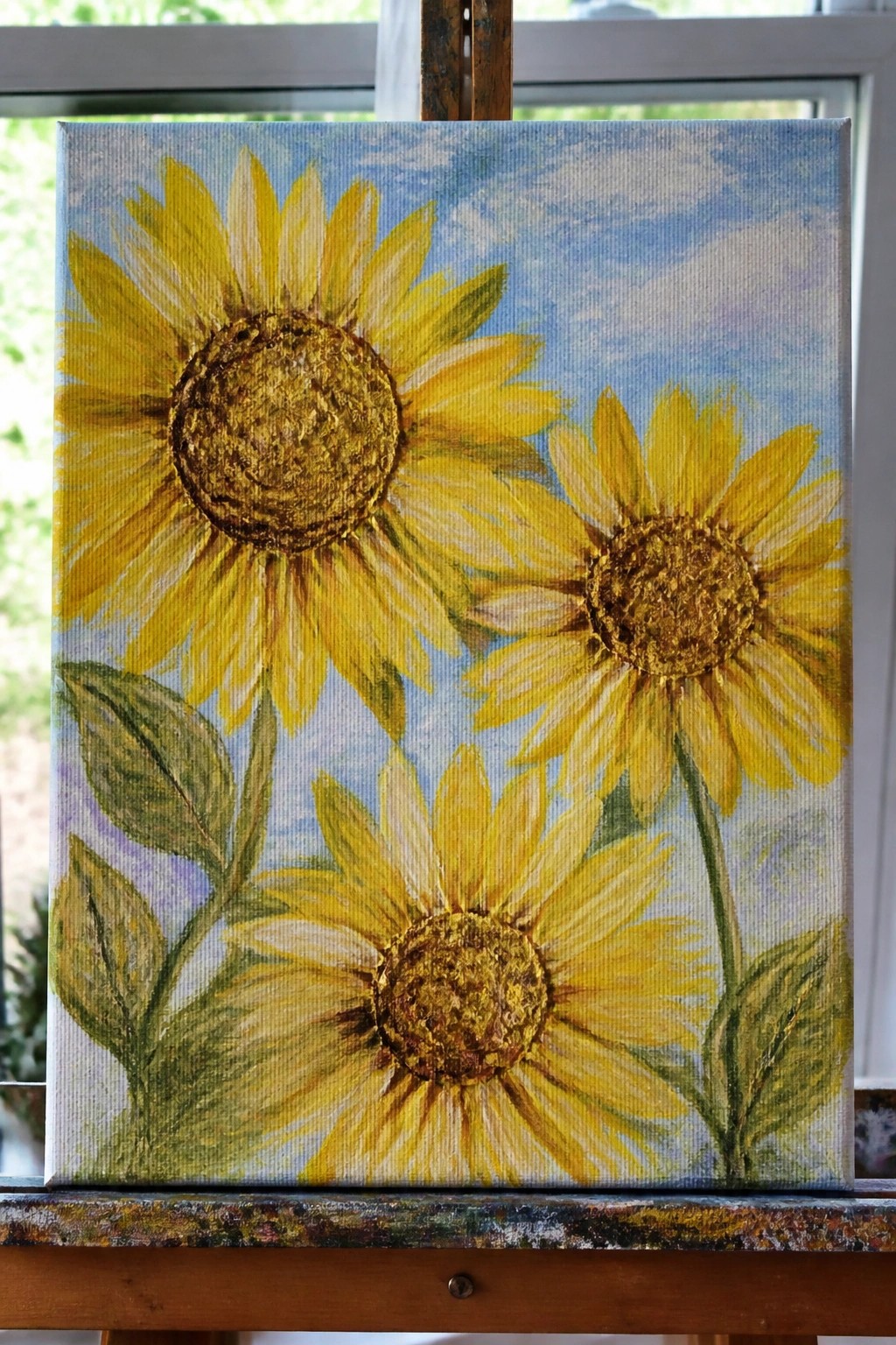 Three yellow sunflowers with green leaves on blue sky painted on canvas atop wooden easel