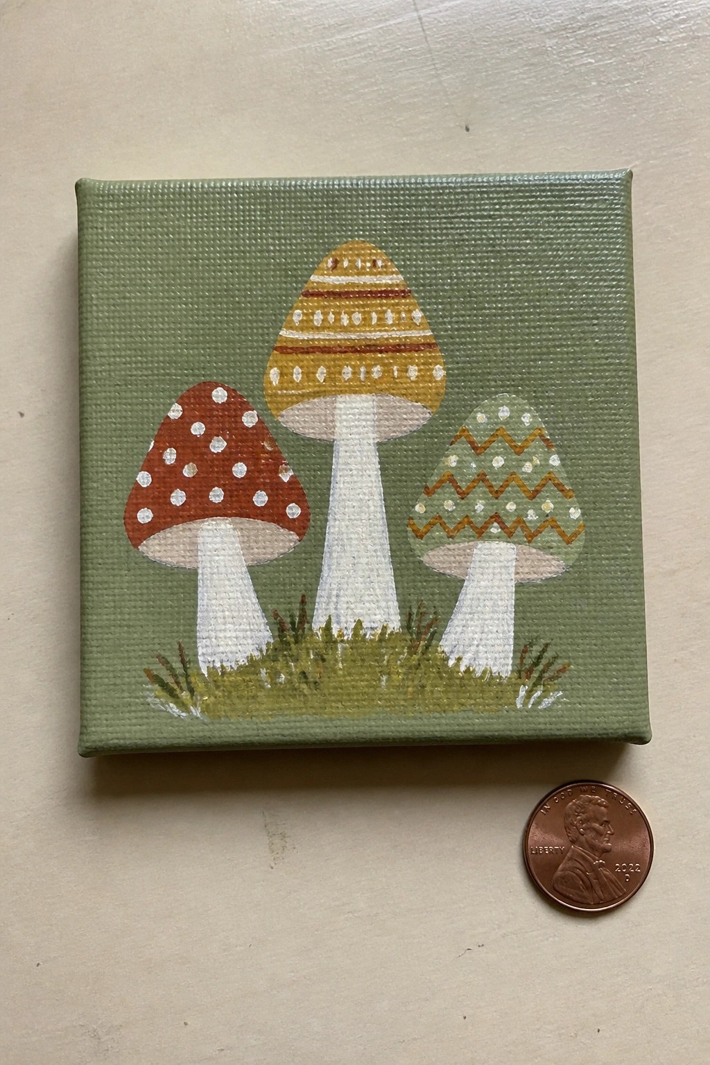 Three patterned mushrooms on small green canvas beside a US penny.