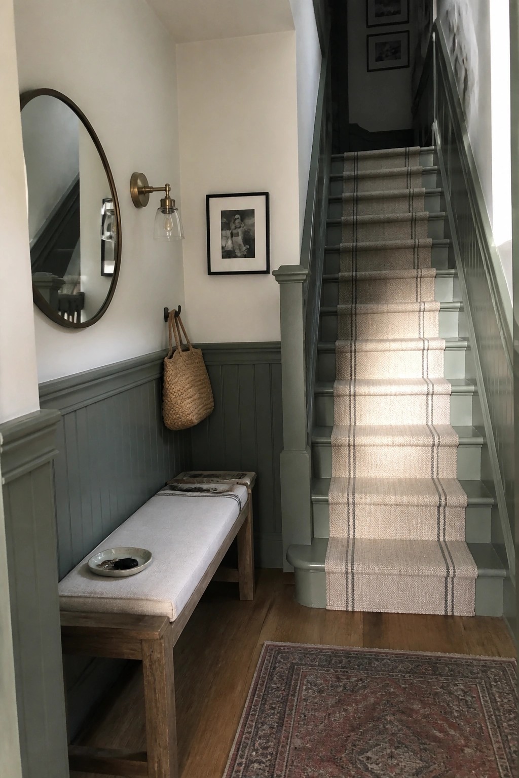 Gray-green wainscoting and stair trim in a narrow home entry with wood flooring and a bench.