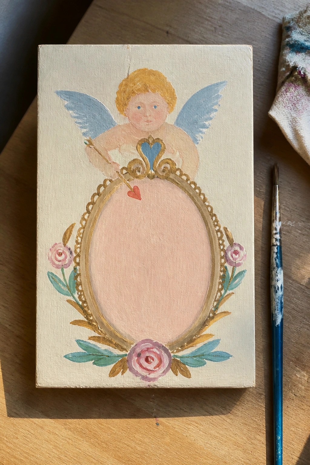 Painted canvas of cherub with blue wings above ornate gold oval frame with pink roses