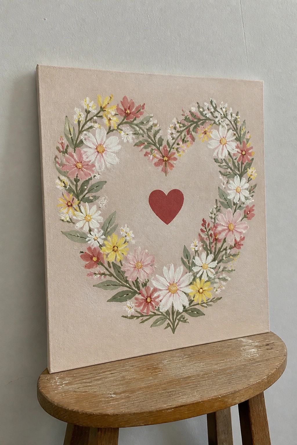 Heart-shaped floral wreath painting with pink, white, yellow flowers and red heart on canvas resting on wooden stool