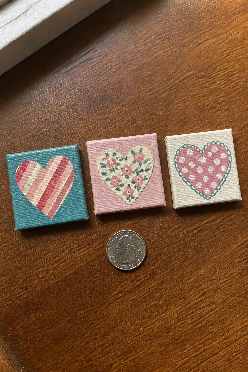Three miniature canvases with patterned hearts beside a quarter on wooden table