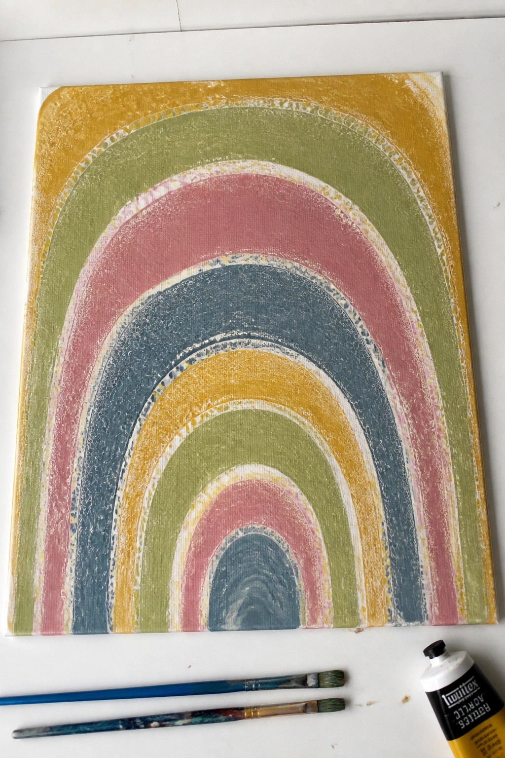 Colorful textured rainbow painting on canvas with paintbrushes and yellow acrylic paint tube.