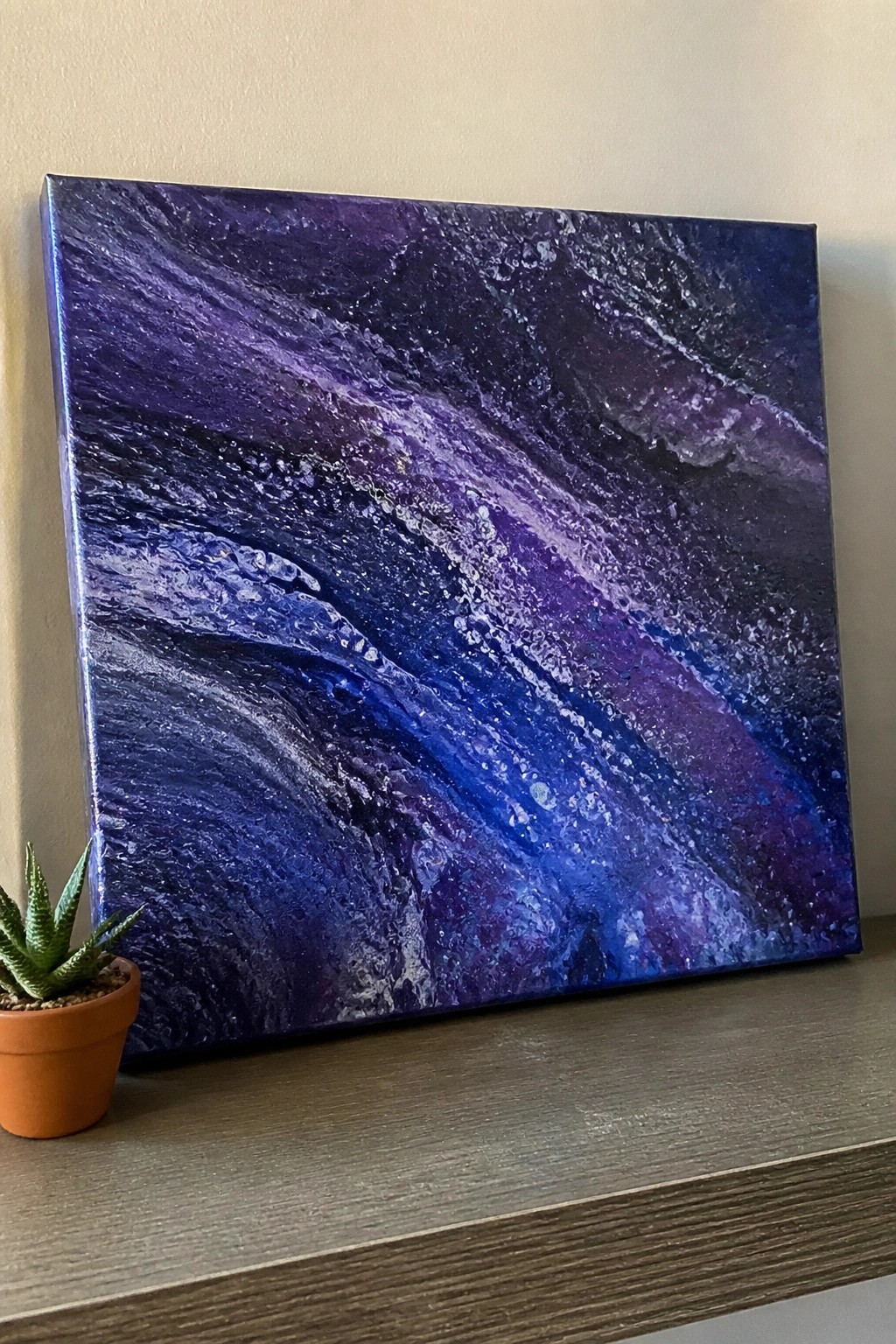 Abstract purple-blue swirled painting on canvas beside potted succulent on wooden shelf
