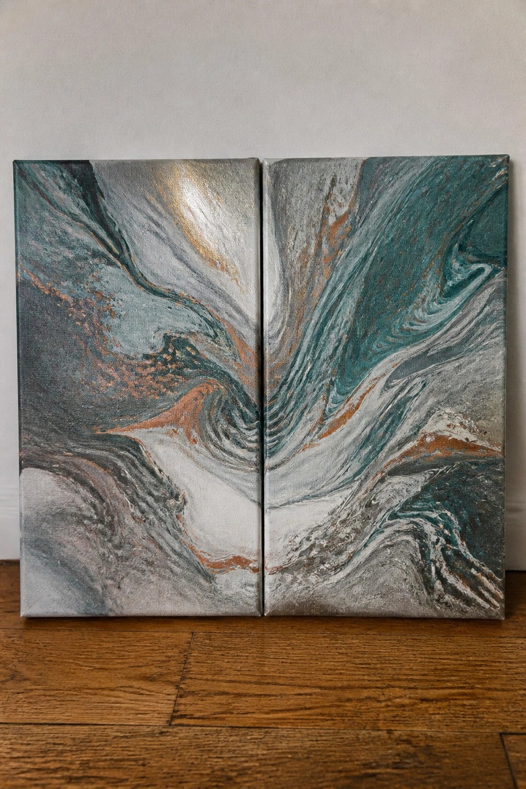 Two abstract fluid paintings with swirling teal, gray, and copper on wooden floor