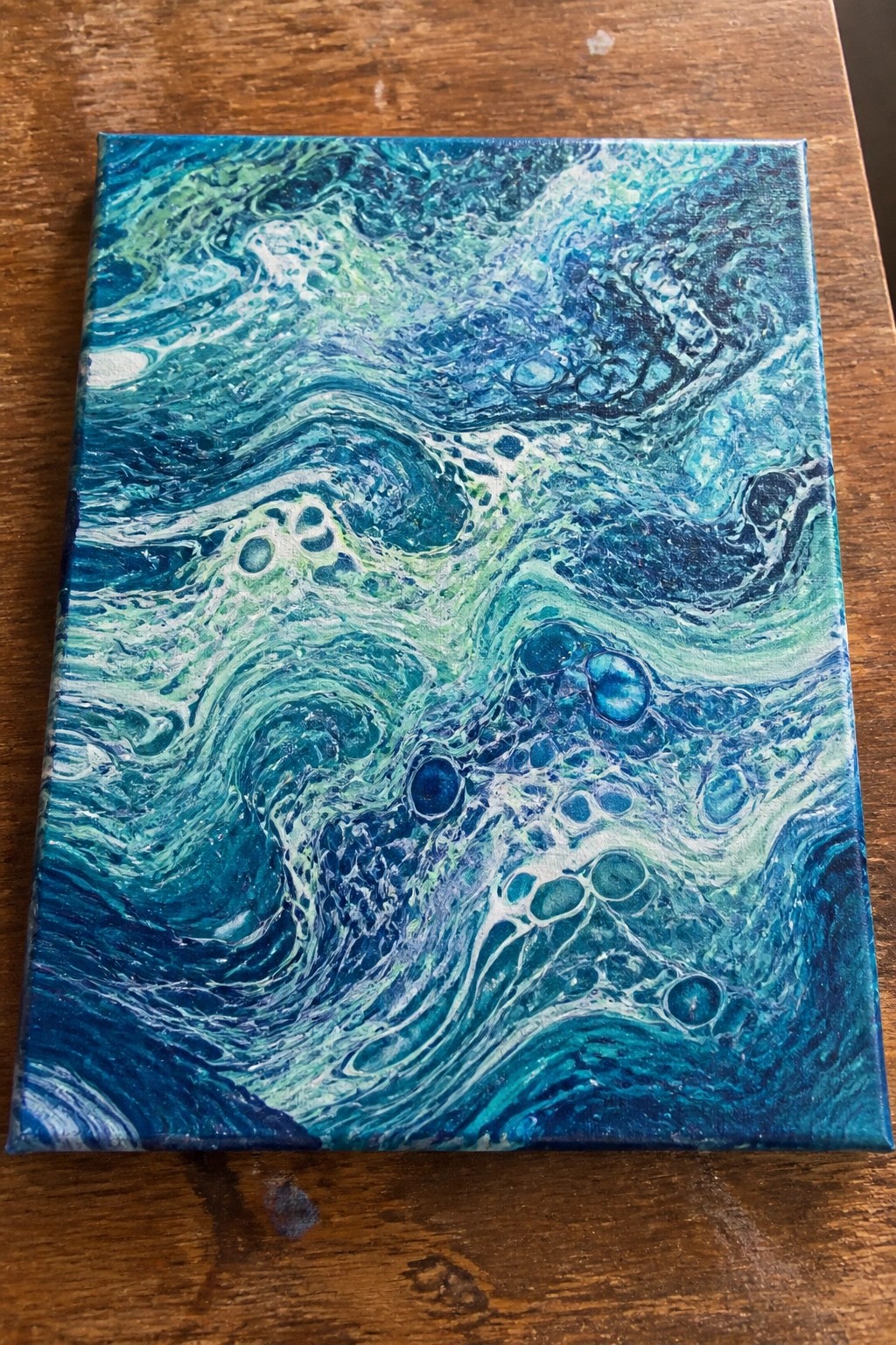 Abstract fluid art canvas with swirling blues, greens, and bubbles on wood.