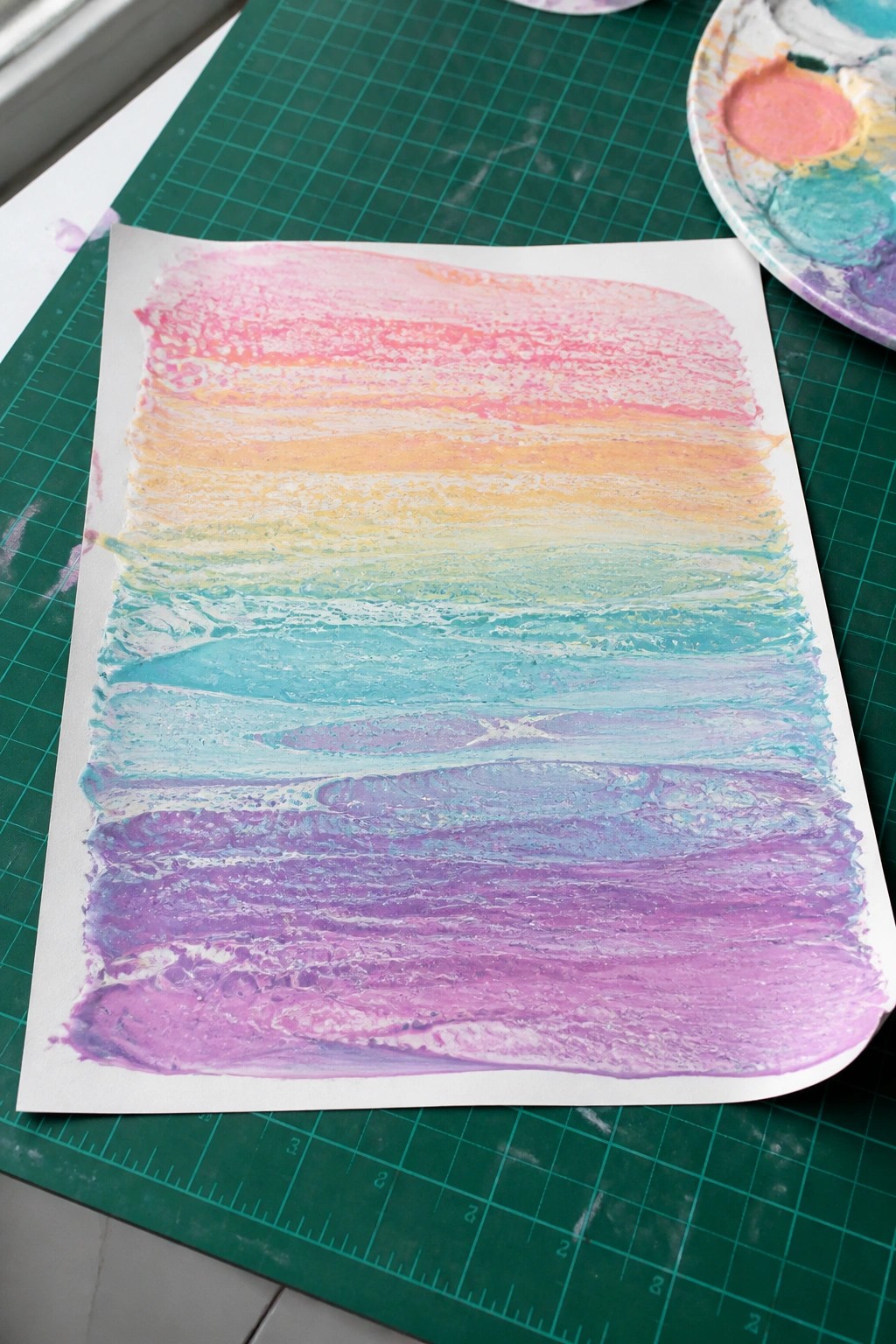 Rainbow paint in thick horizontal stripes on paper over green cutting mat
