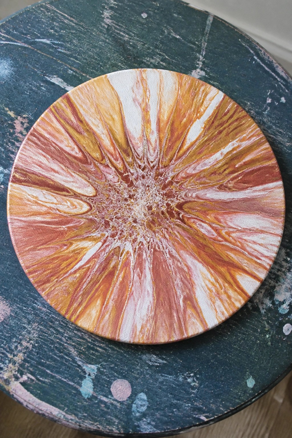 Abstract fluid art in orange, red, and white on round canvas atop green table