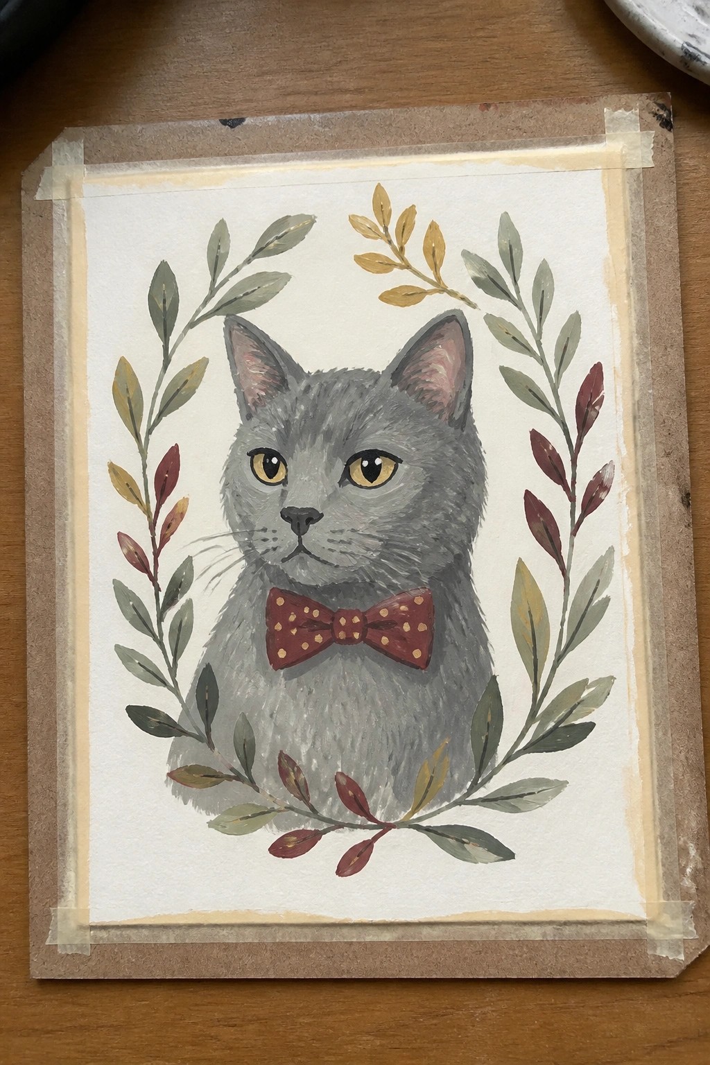 Gray cat in maroon polka-dot bow tie amid watercolor autumn leaves on taped paper