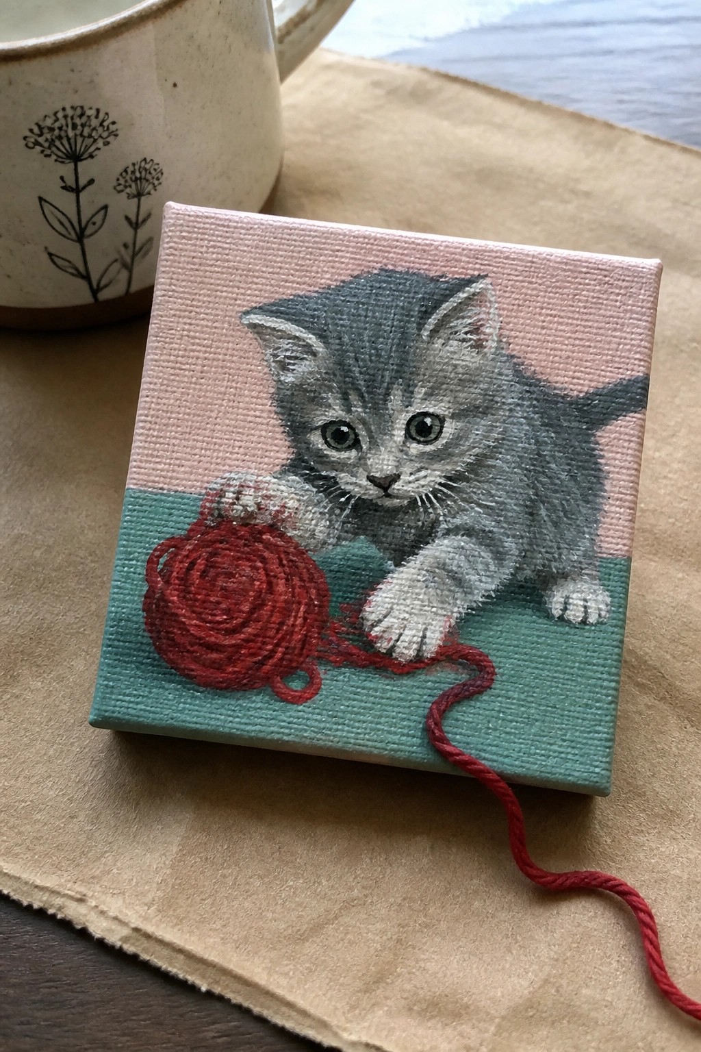 Miniature canvas painting of gray kitten playing with red yarn ball beside mug