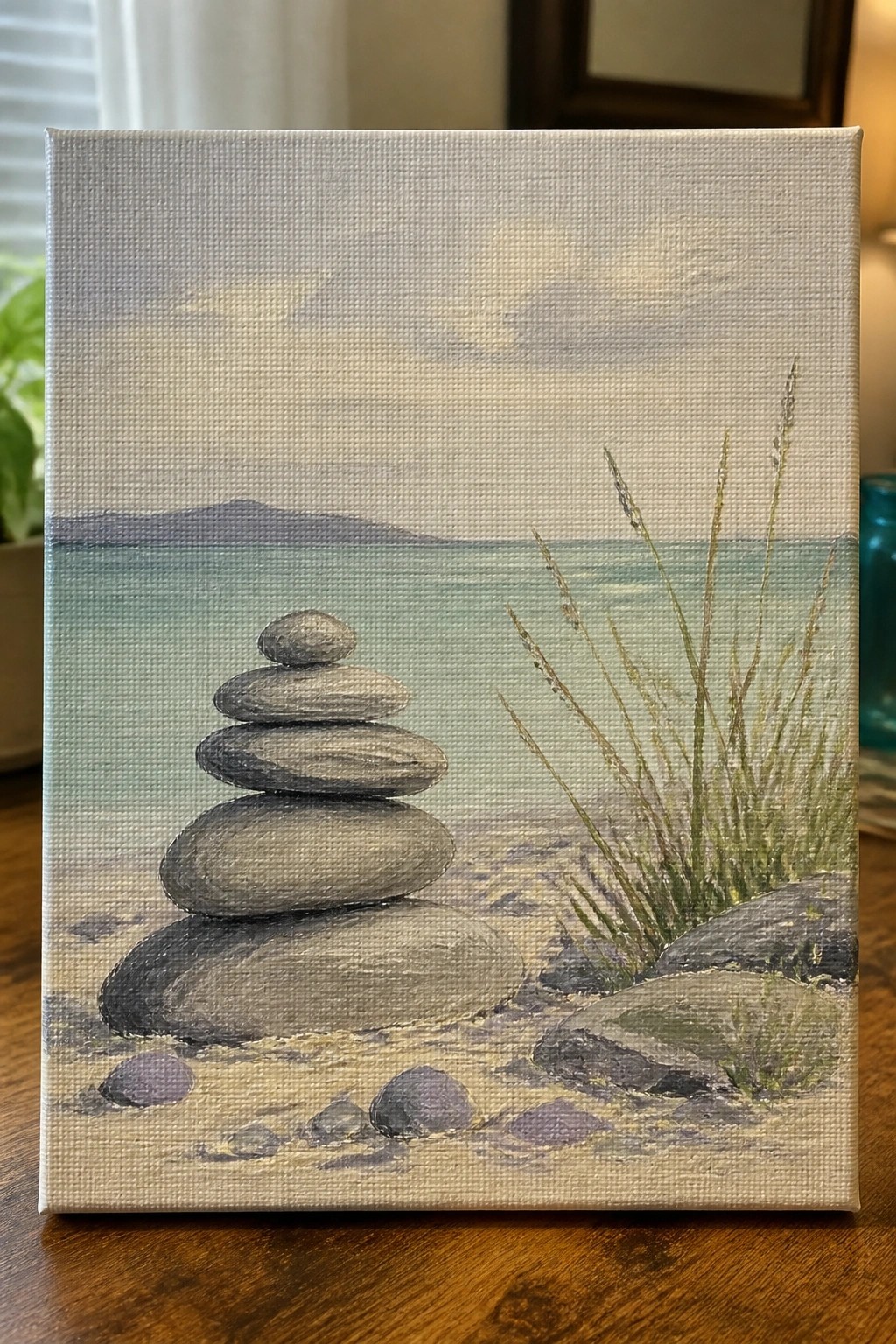 Stacked stones on sandy beach by calm water with distant hills and tall grass.