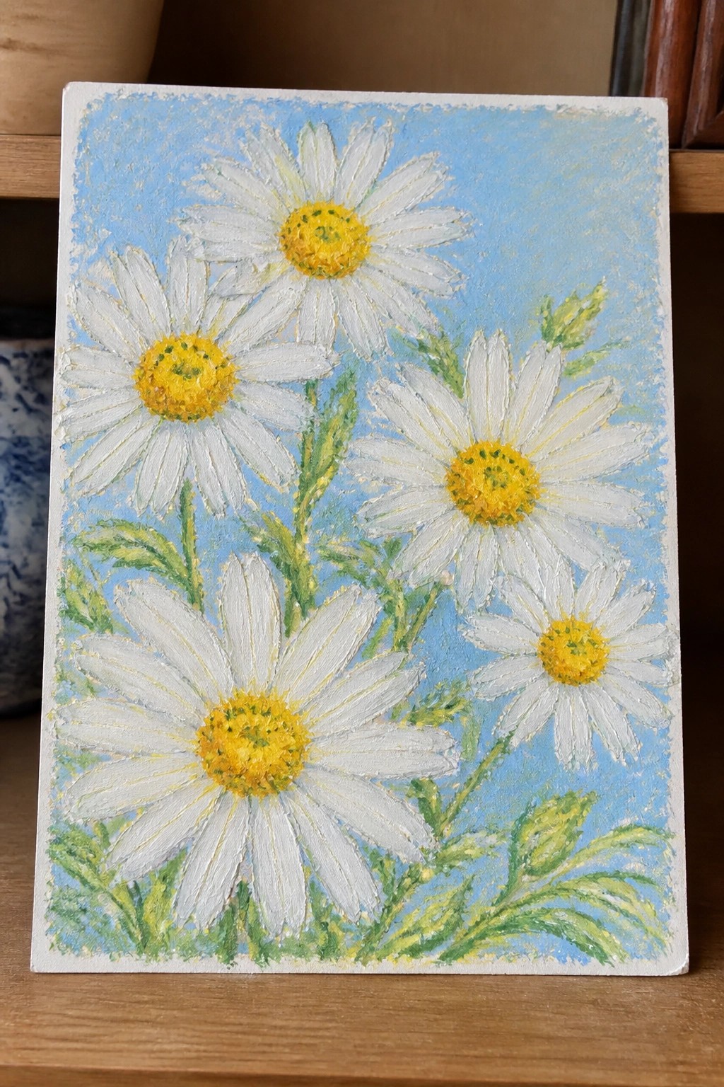 Painting of white daisies with yellow centers, green leaves on blue background.