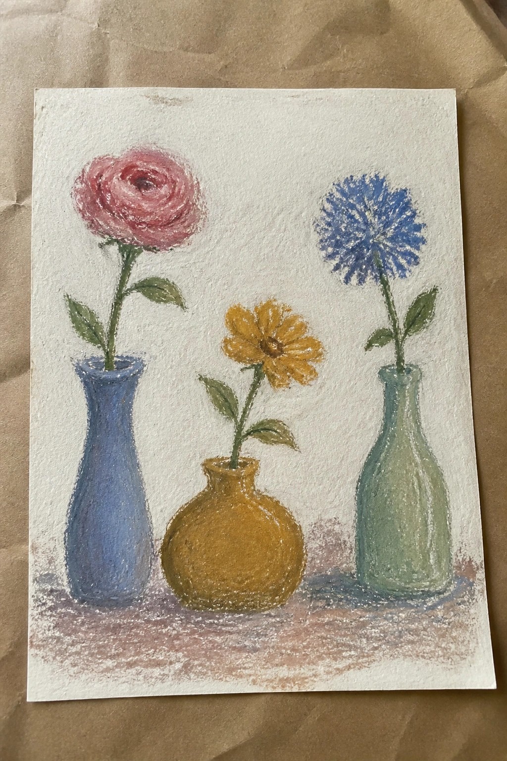Pastel drawing of pink rose in blue vase, yellow bloom in brown, blue flower in green.
