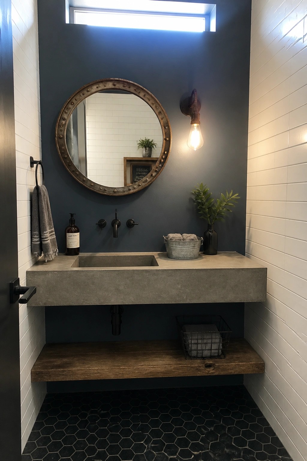 Modern bathroom with floating concrete sink, round riveted mirror, dark blue wall, and black hex tiles.