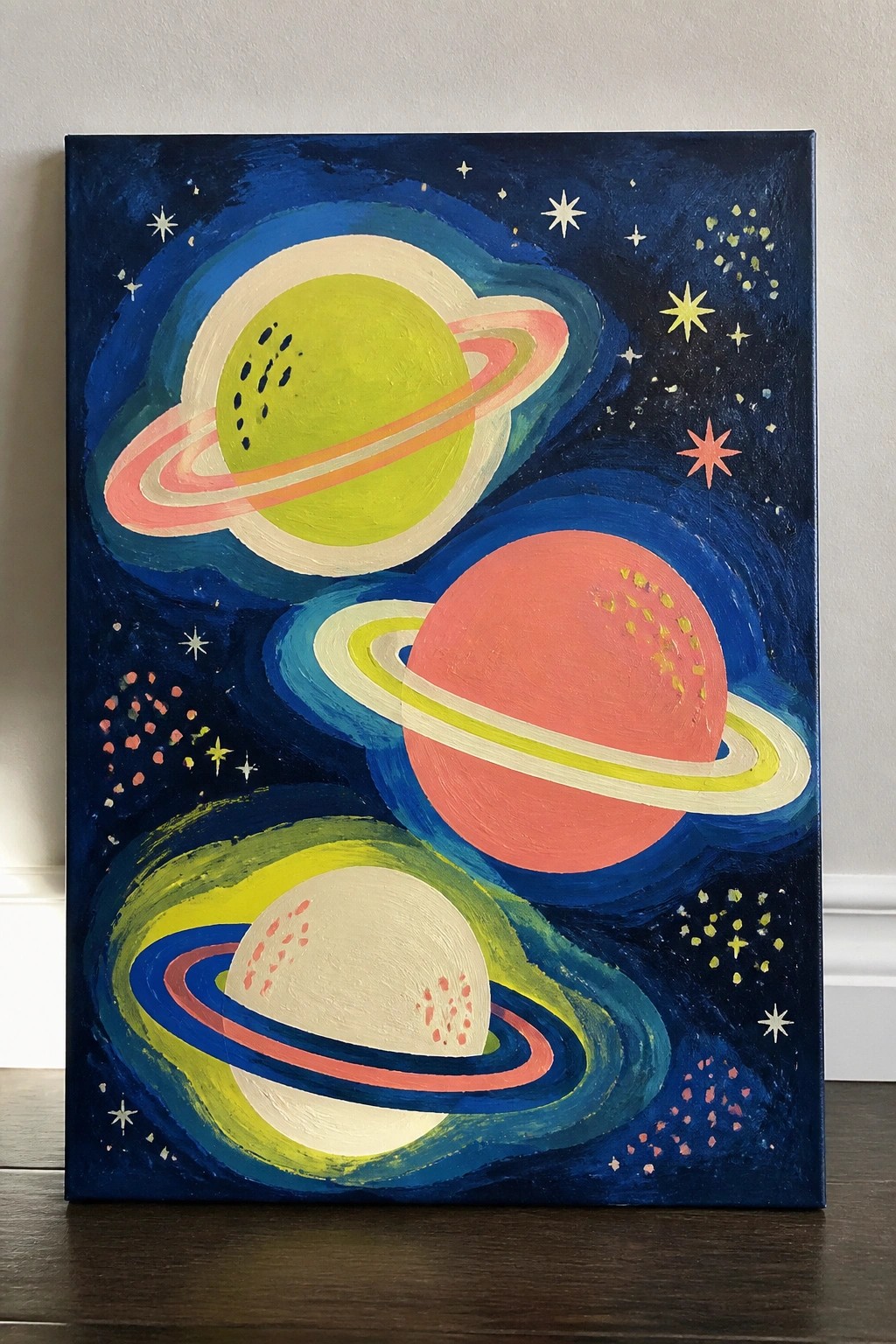 Vibrant painting of three colorful ringed planets against a dark blue starry sky.
