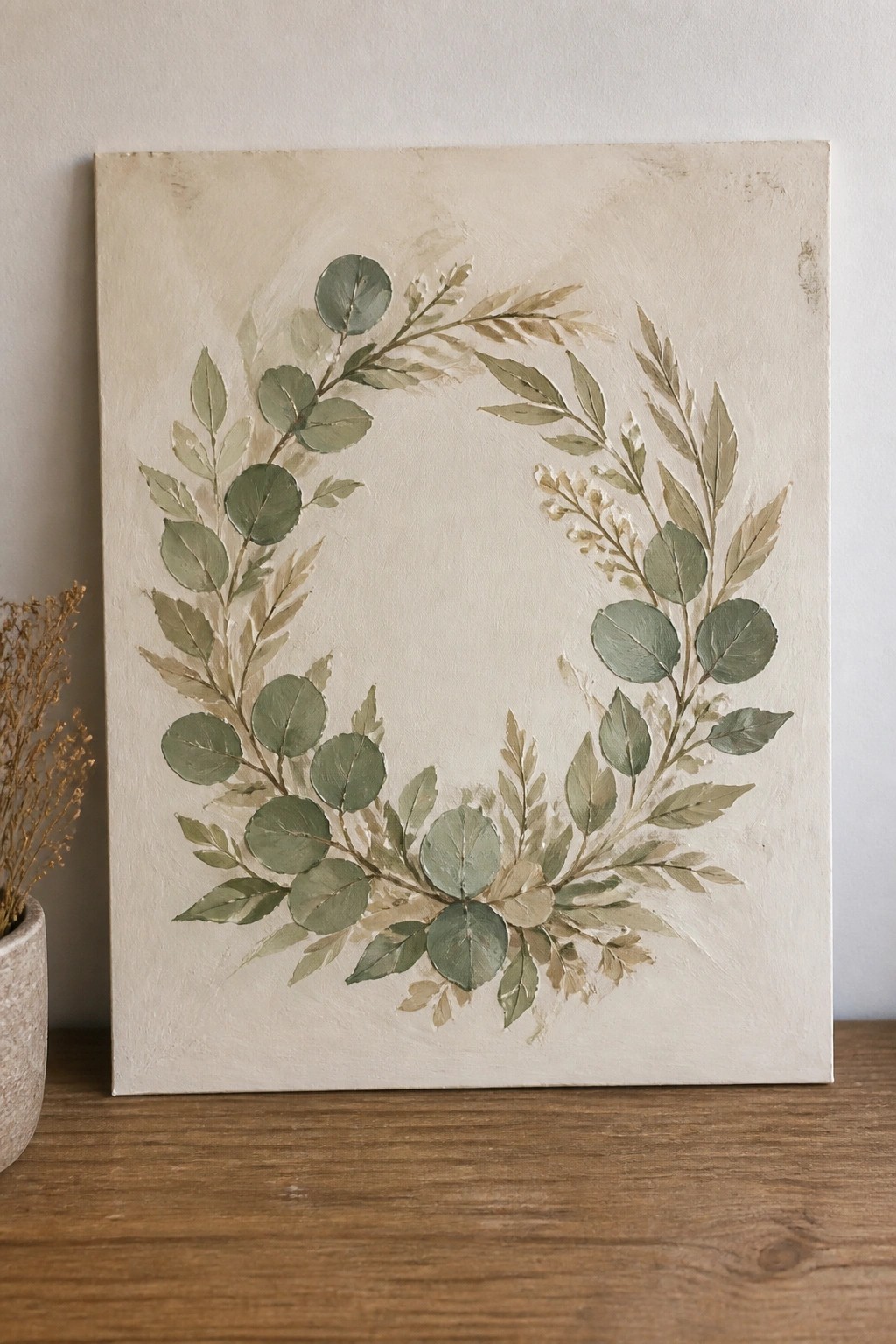 Canvas with painted green leaf wreath on wooden table beside ceramic vase