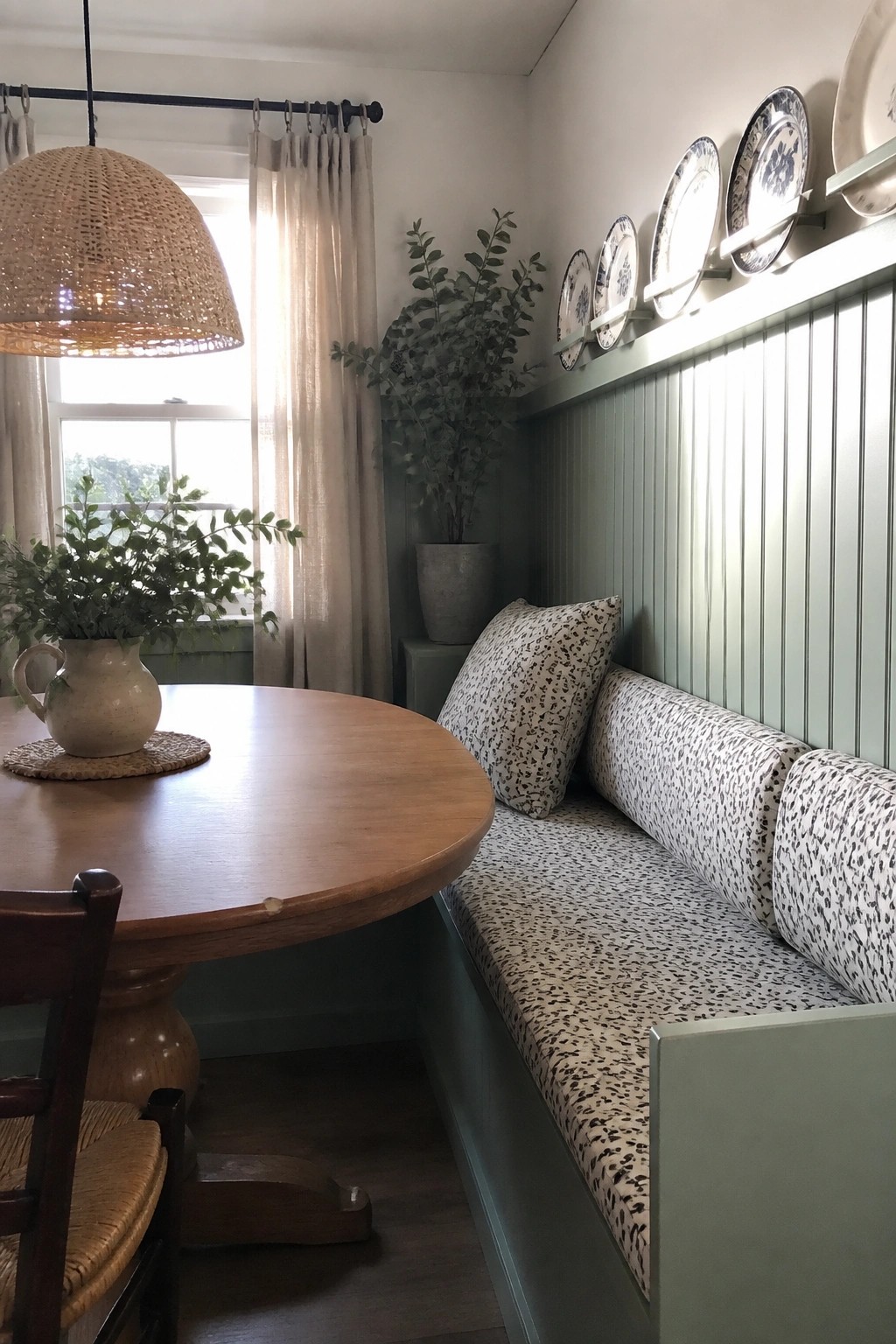 Cozy dining nook with round wooden table, green banquette, patterned cushions, and woven pendant light.