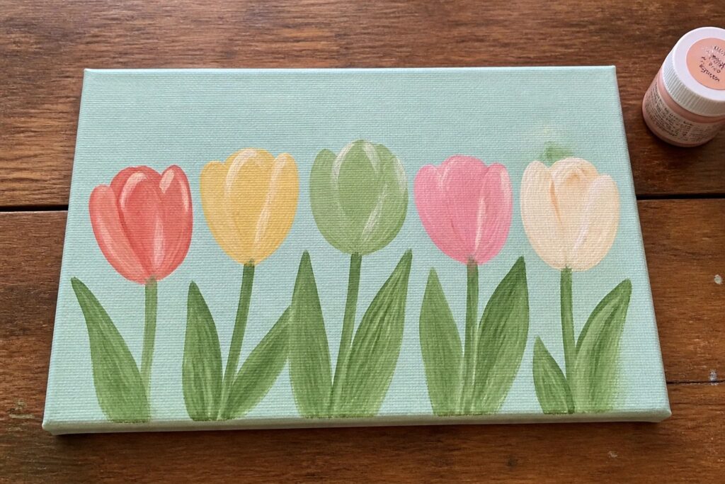 A canvas with five tulips painted in red-orange, yellow, green, pink, and beige on a light green background, next to a bottle of Golden light green acrylic paint.
