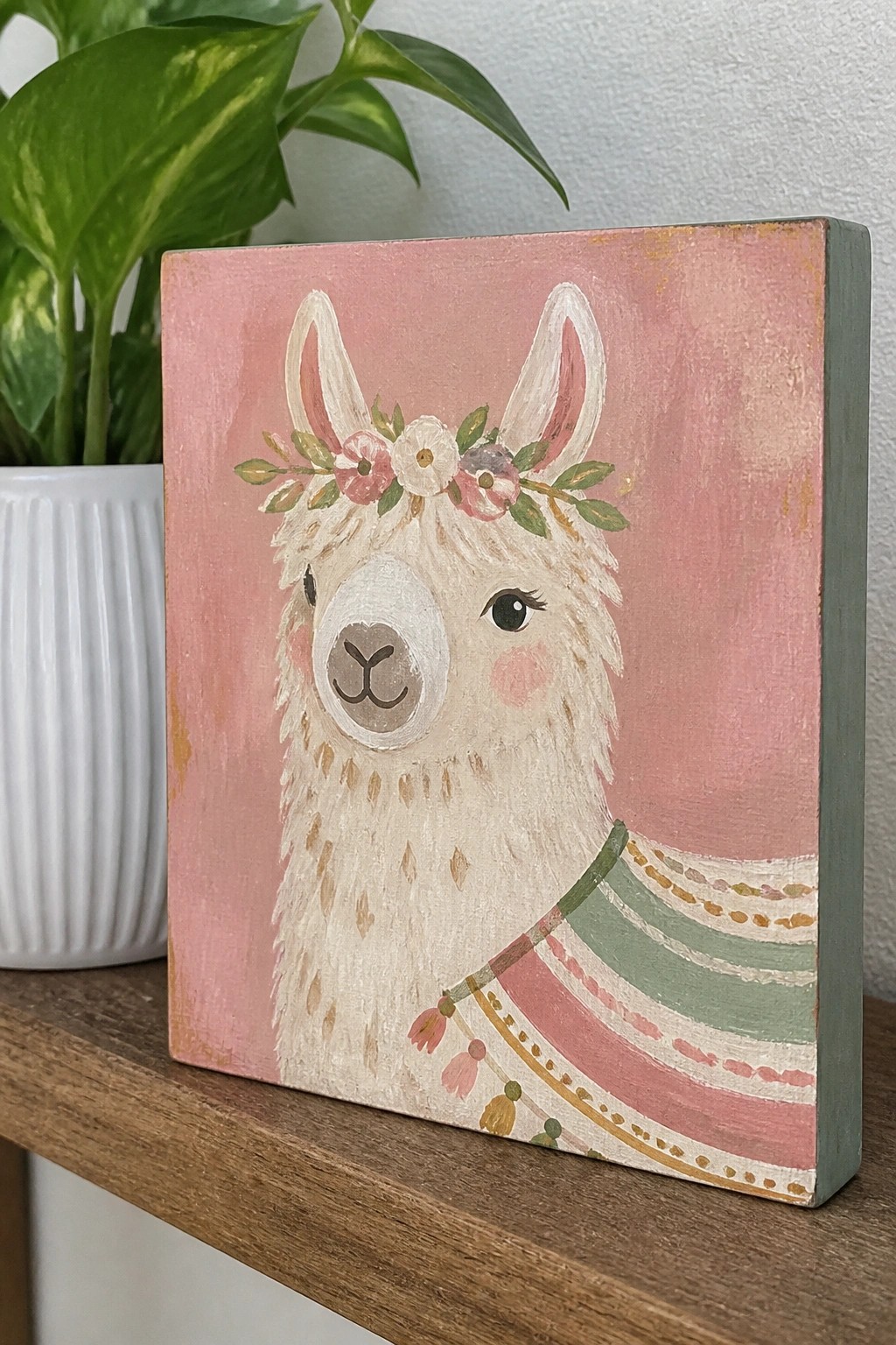 Cute llama painting with floral crown and colorful blanket on pink canvas beside plant.