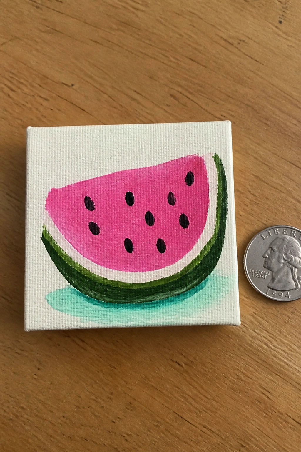 Mini canvas painting of pink watermelon slice beside 1994 quarter on wood.