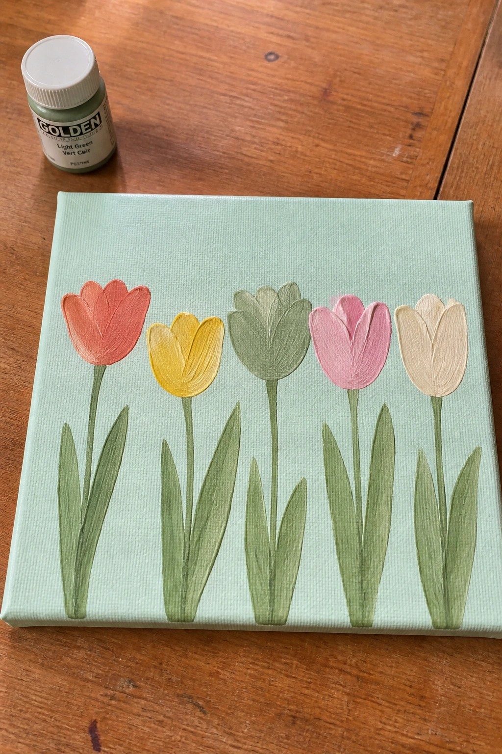 A canvas with five tulips painted in red-orange, yellow, green, pink, and beige on a light green background, next to a bottle of Golden light green acrylic paint.