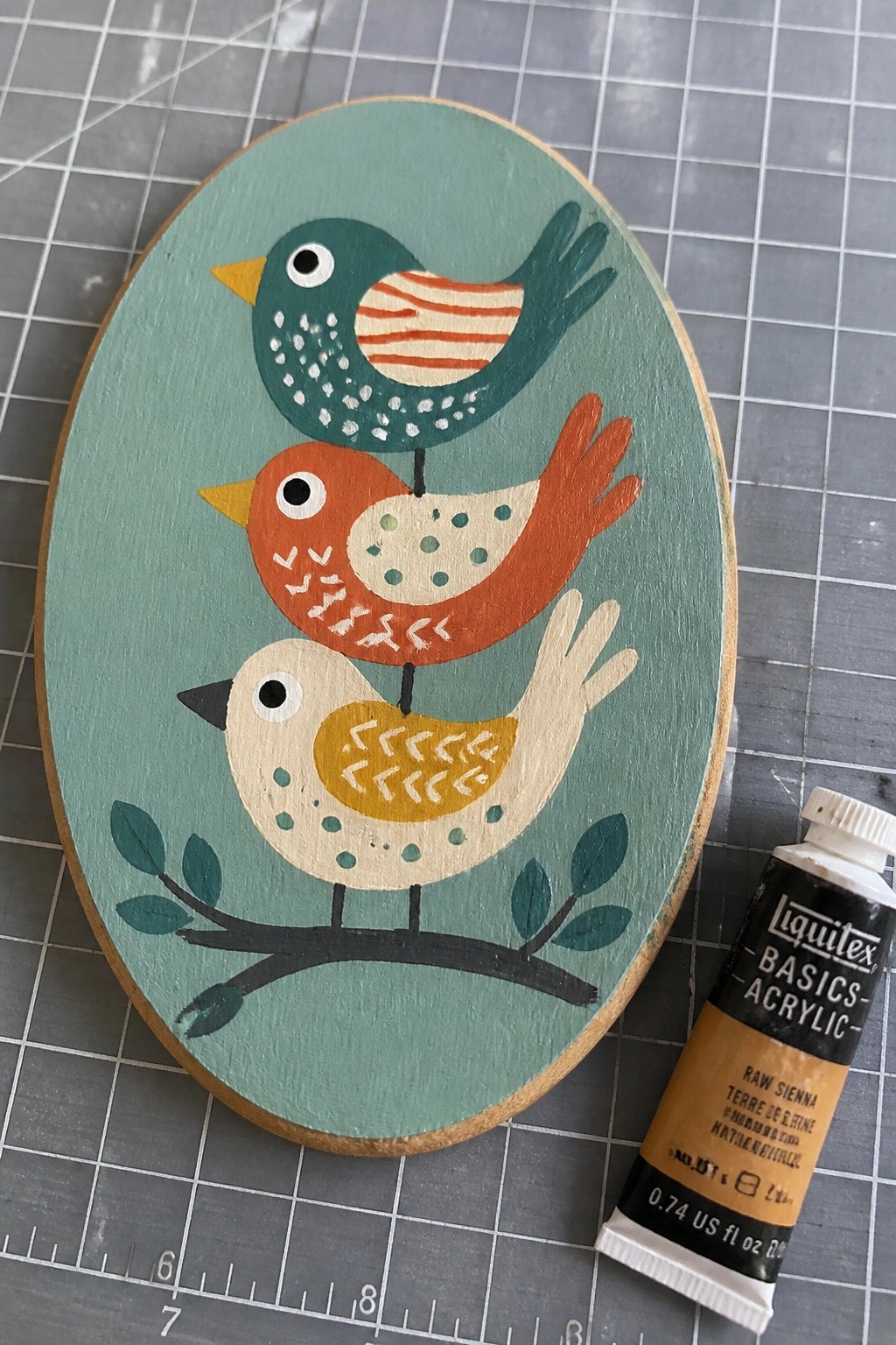 Oval teal plaque with three stacked colorful folk-art birds beside raw sienna paint tube