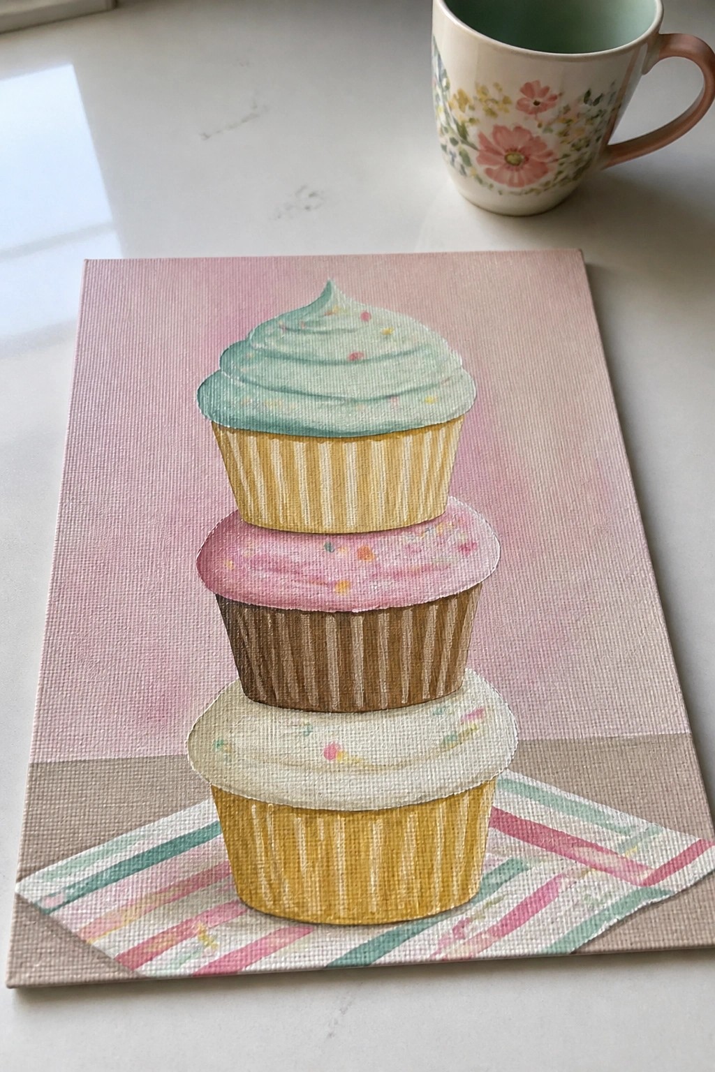 Watercolor painting of three stacked cupcakes with pastel frosting beside floral mug.