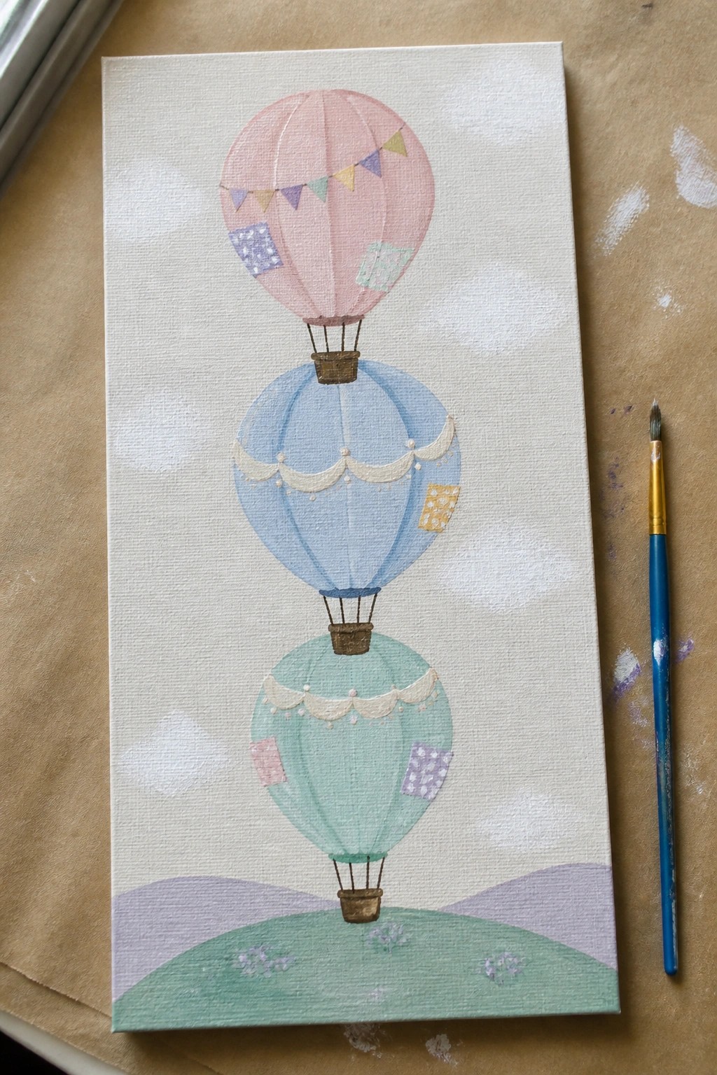 Three stacked pastel hot air balloons over green hills on canvas with paintbrush