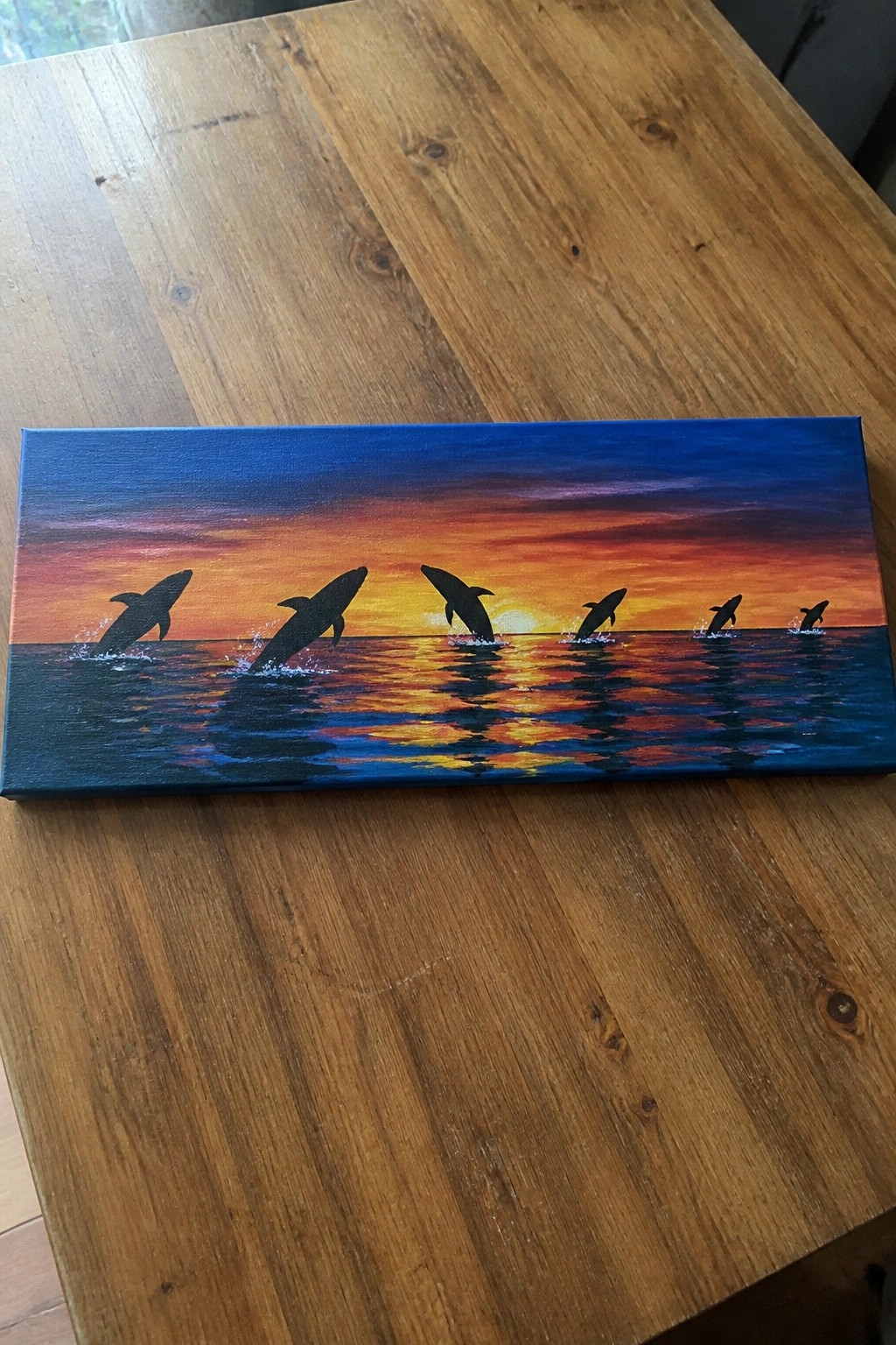 A rectangular canvas on a wooden table shows six black dolphin silhouettes jumping from the ocean against a vibrant orange and blue sunset with reflections in the water.