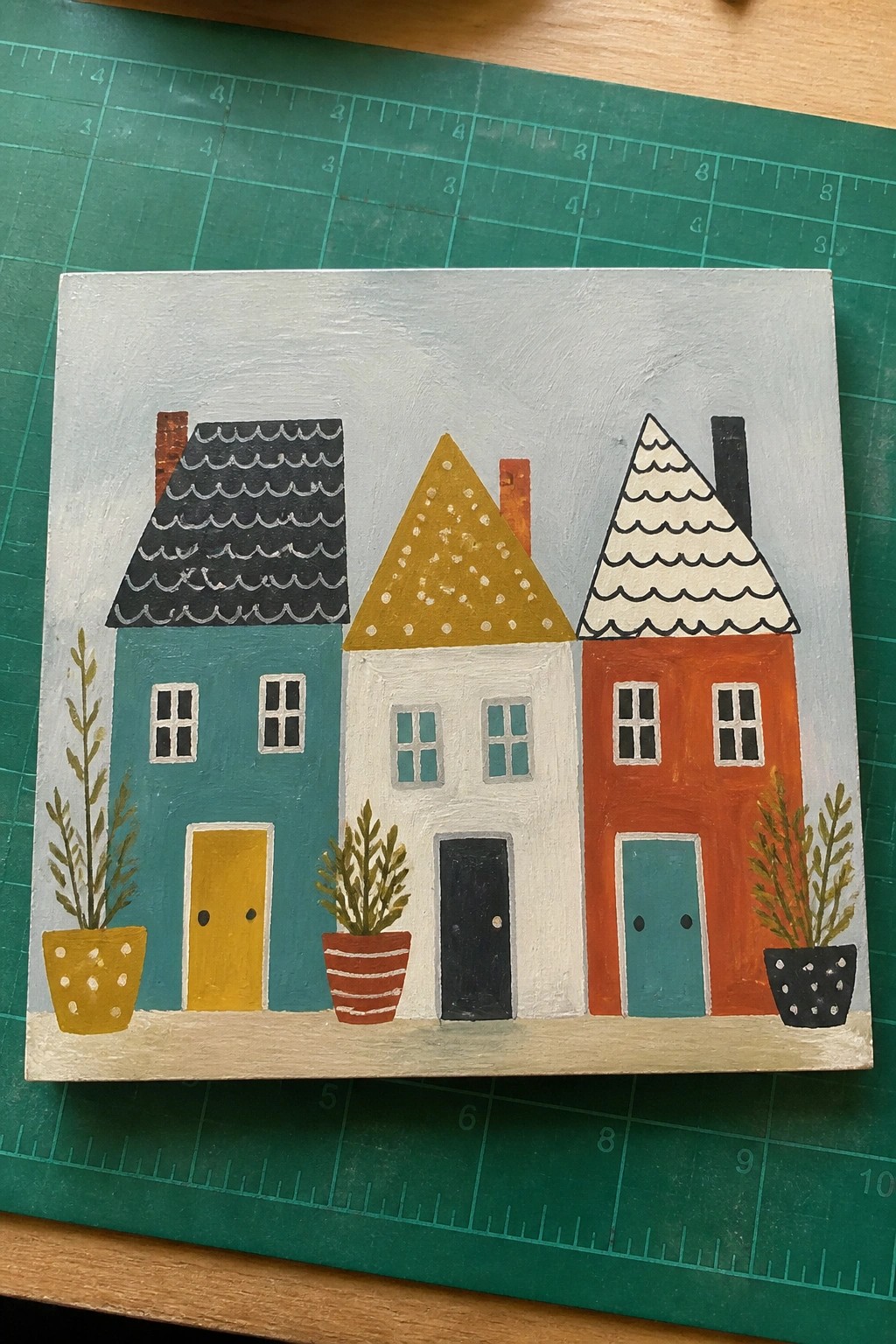 Colorful folk-art painting of three houses with patterned roofs and potted plants