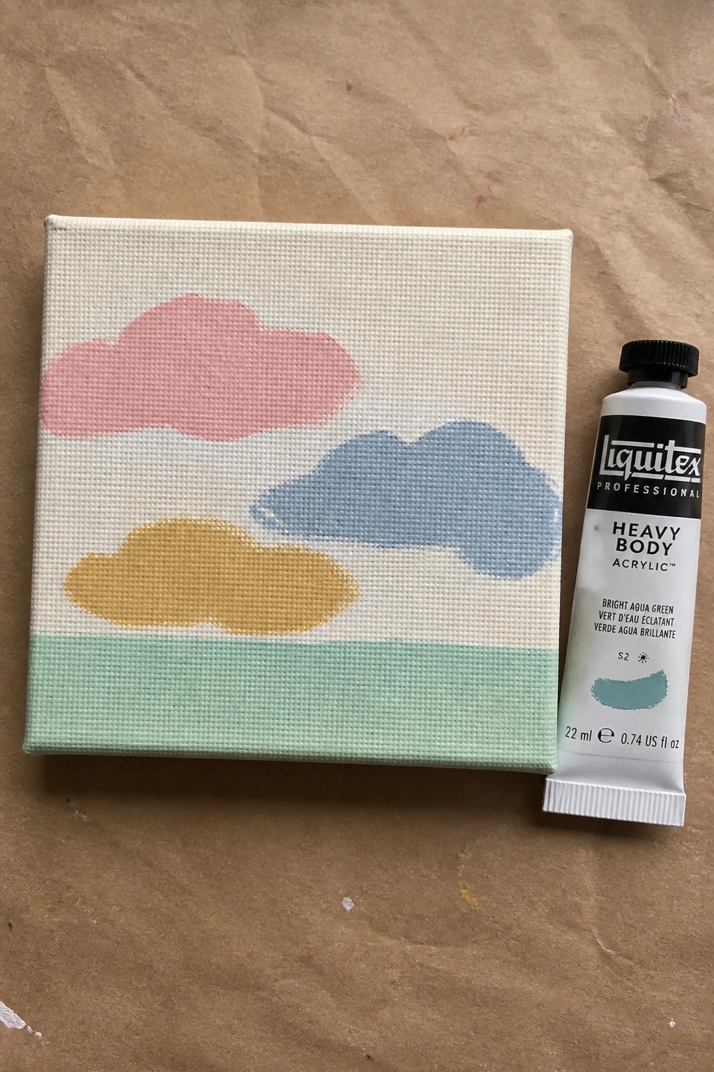 Small canvas with pink, blue, yellow clouds above green stripe beside paint tube.