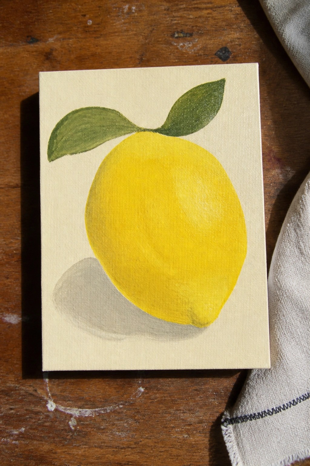 Yellow lemon with green leaves painted on small canvas atop wooden table